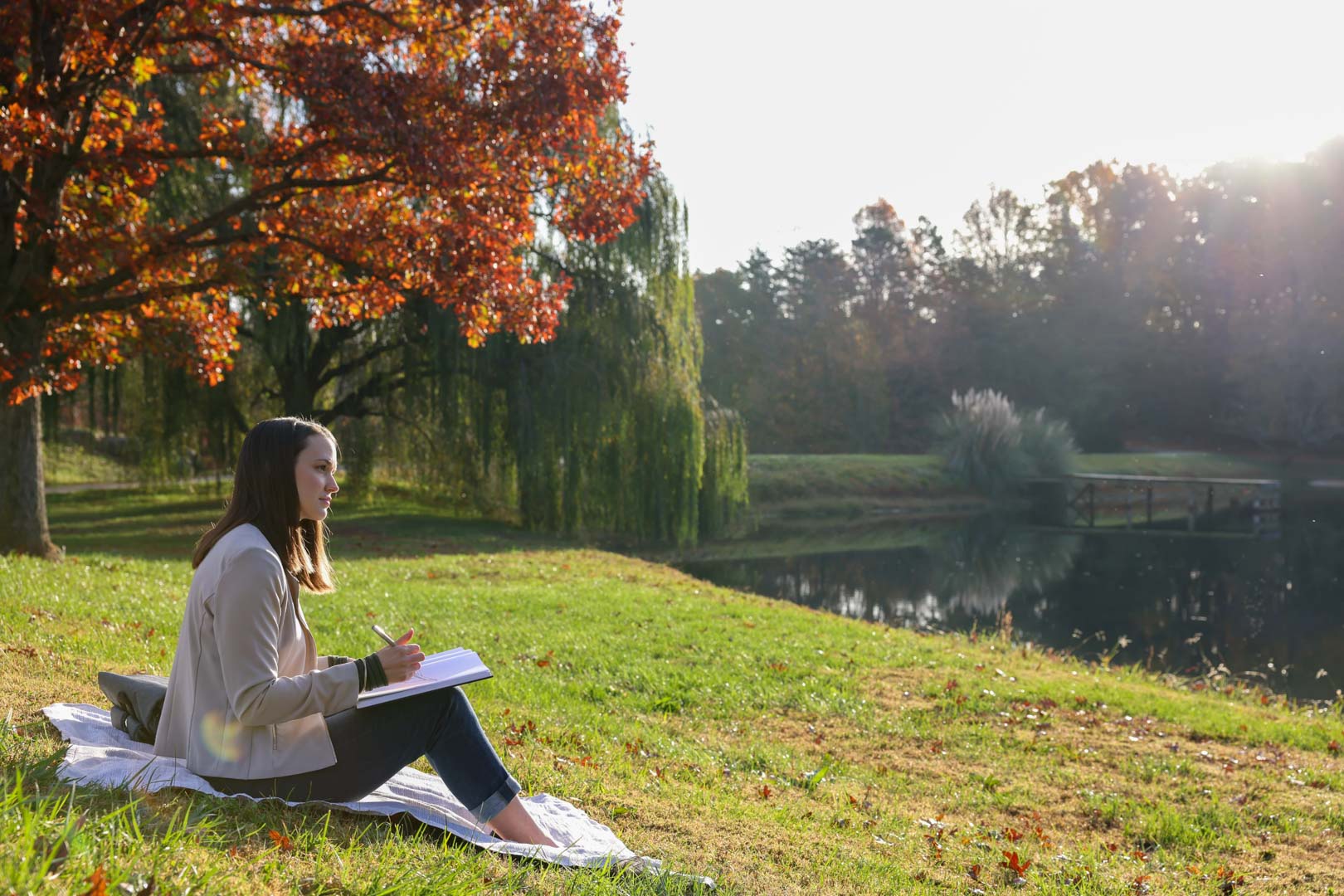 woman sitting in grass with notebook overlooking a park lake
