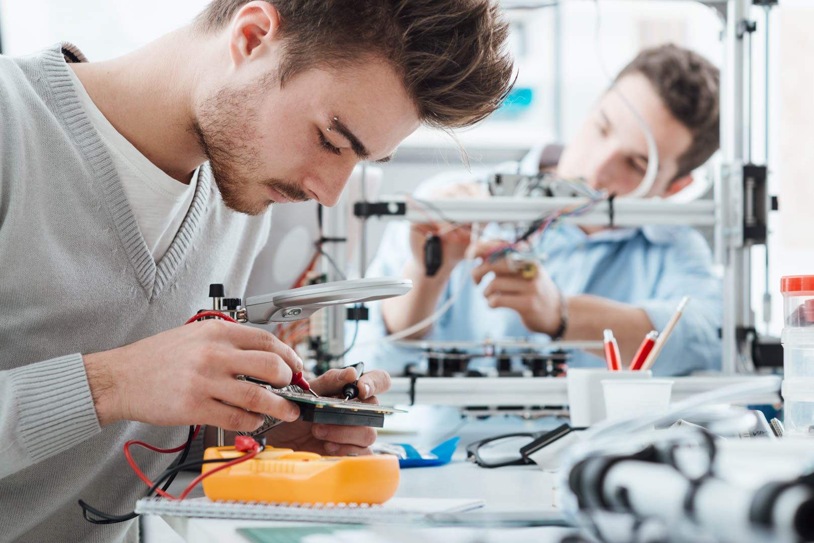 Engineering students working in the lab, a student is using a voltage and current tester, another student in the background is using a 3D printer